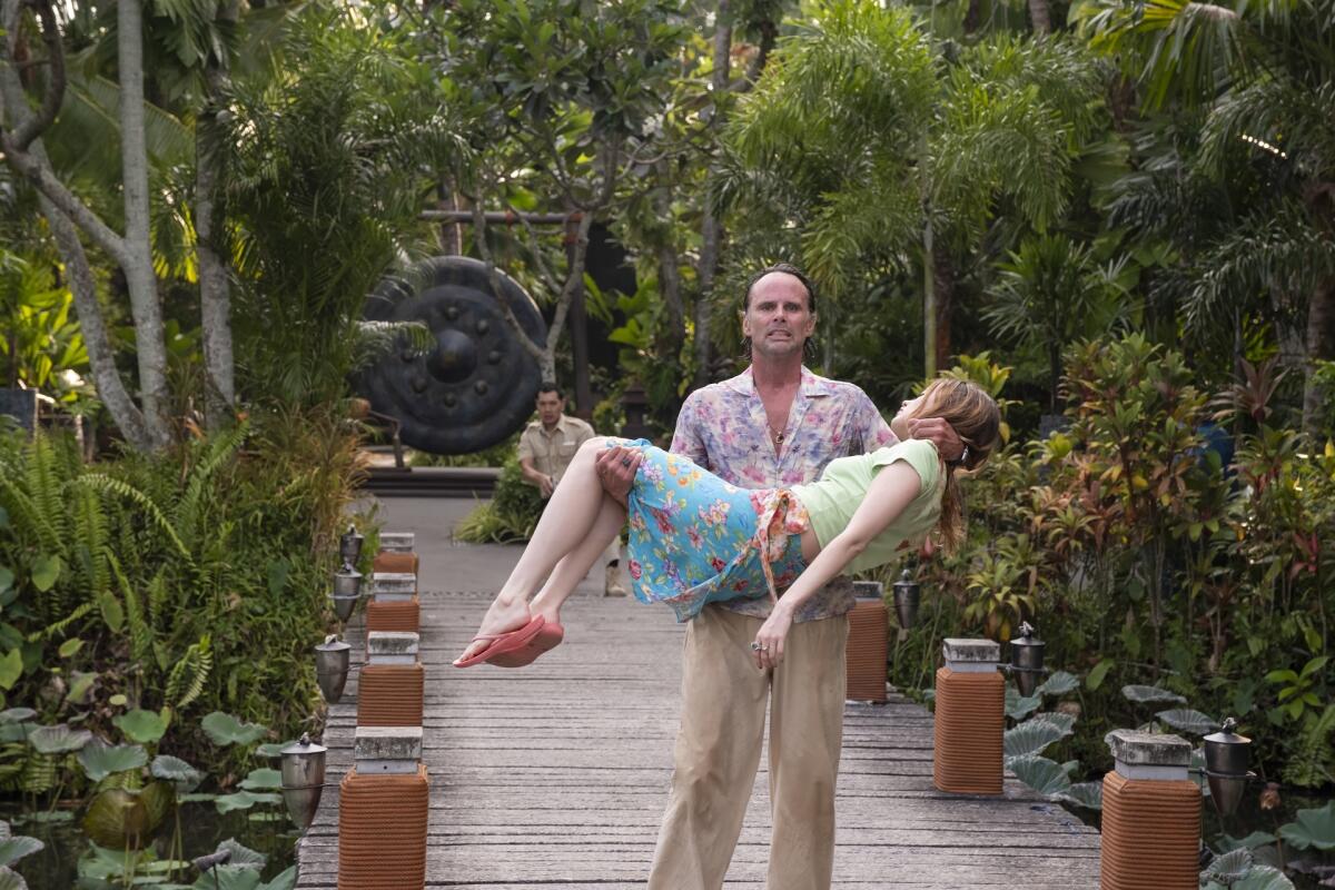 A man carries a woman across a bridge as a man behind him holds and points a gun.