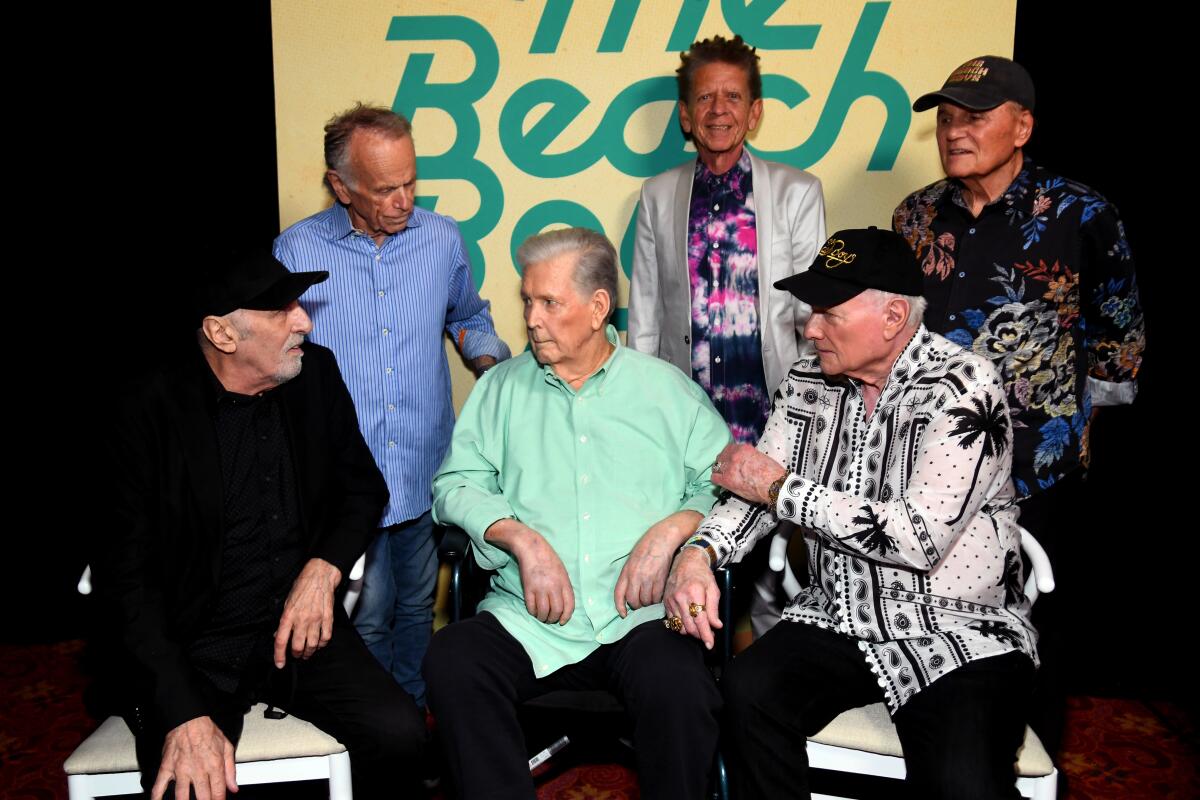 Three Beach Boys sit while three others stand behind them in front of a yellow backdrop with the group's name on it