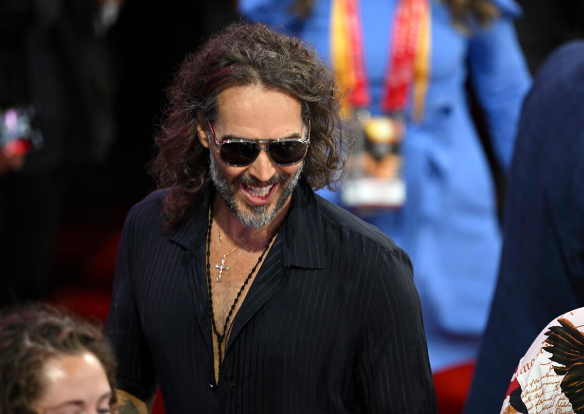 Russell Brand wears dark sunglasses and smiles while wearing a dark unbuttoned shirt baring a necklace with a cross pendant