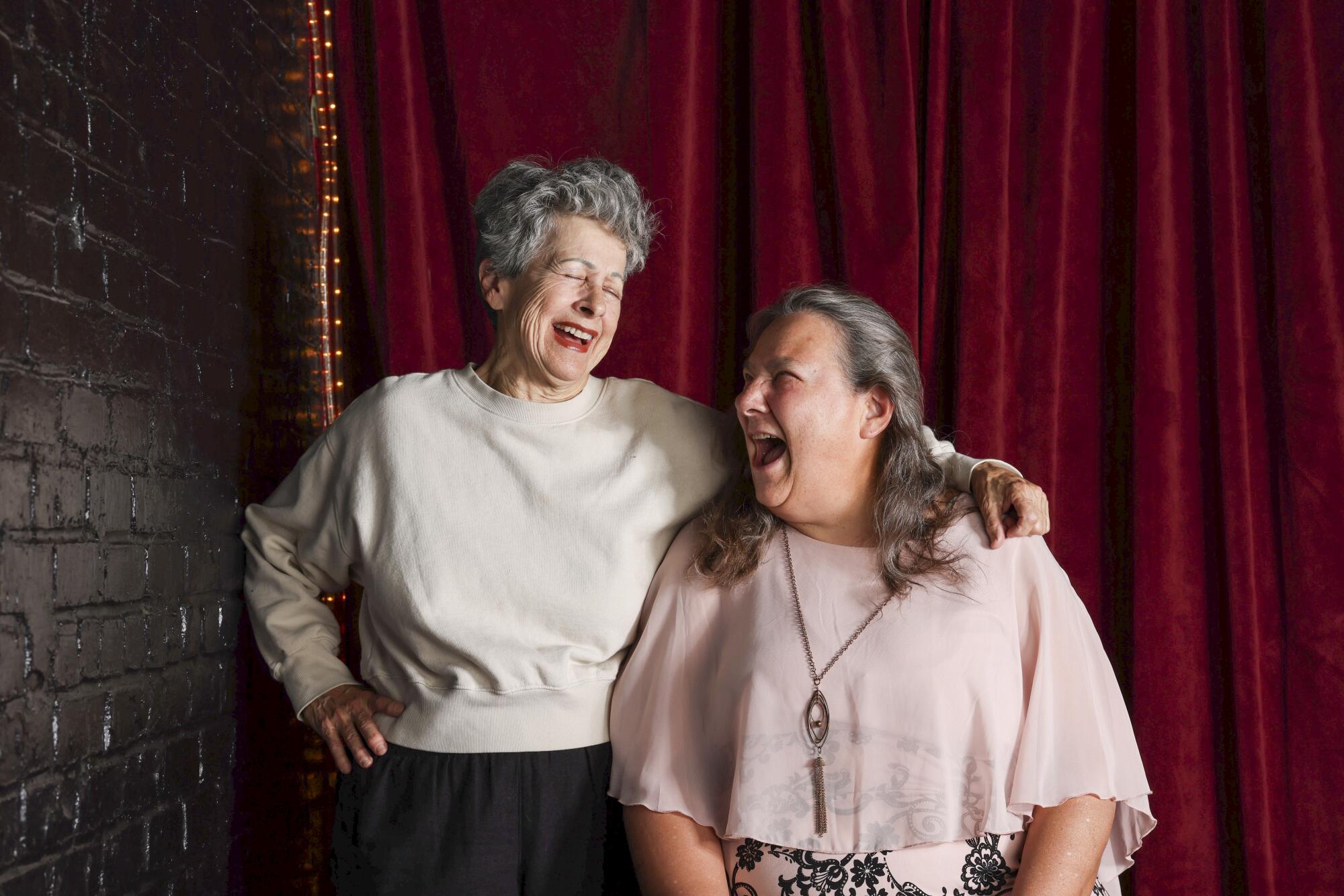 One woman is standing wearing a light-colored top, while the other in a pink top sits. Both are laughing.