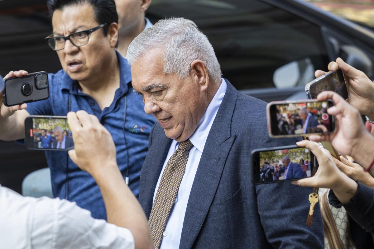 A gray-haired man, in gray suit and brown tie, looks down as others photograph him with their cellphones