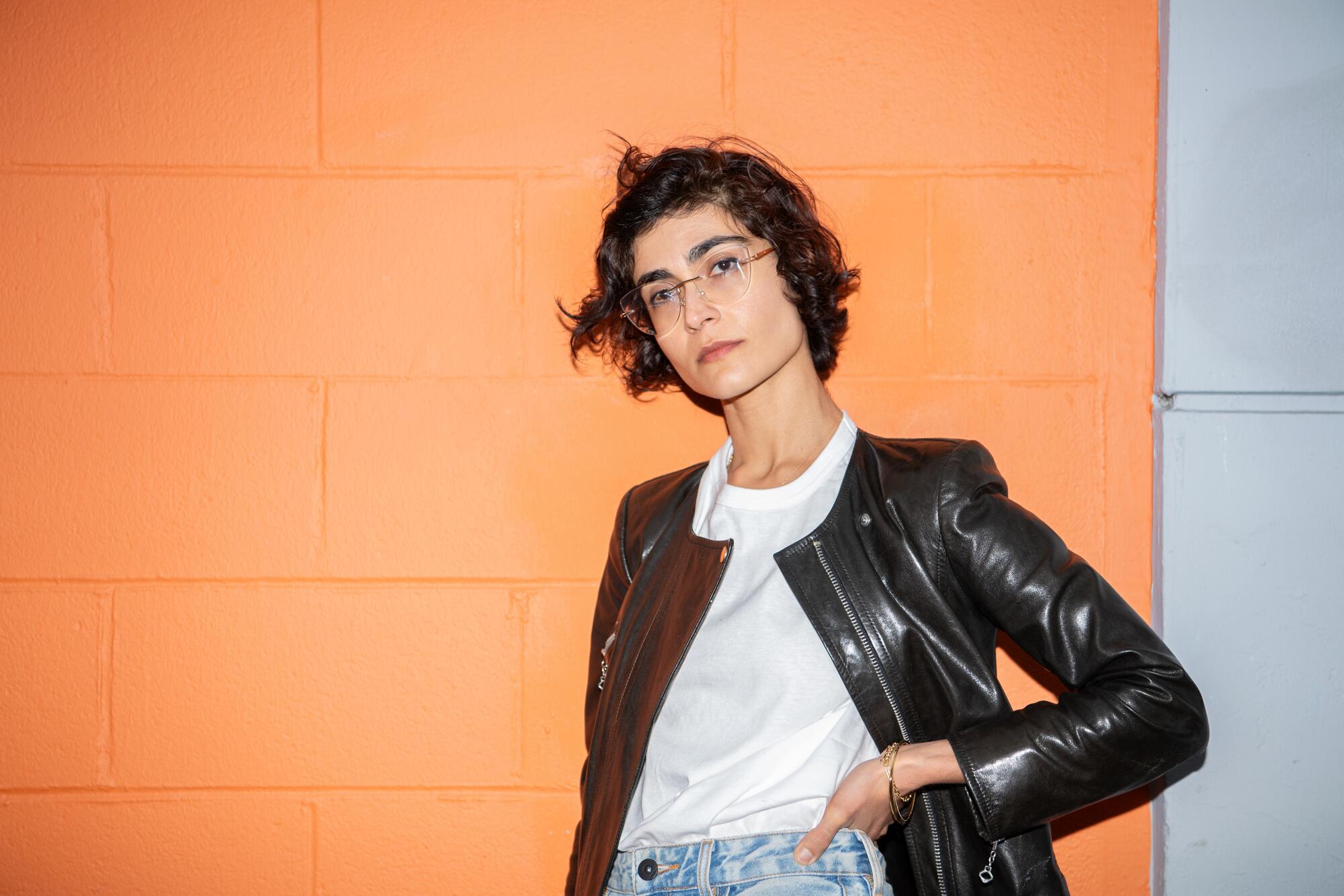 Author Mariam Rahmani poses in a leather jacket in front of an orange wall.