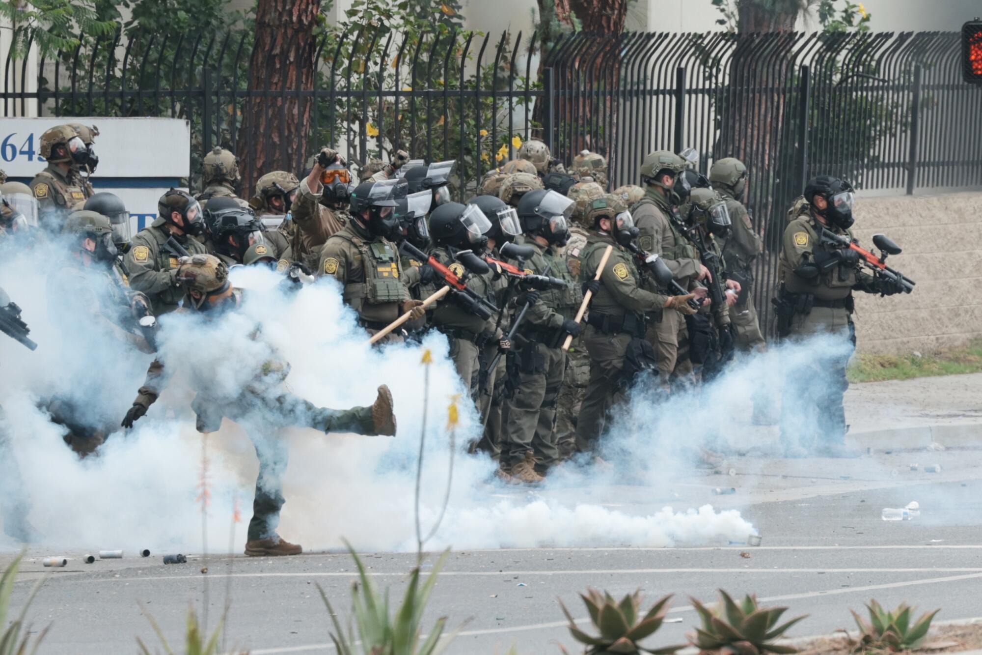 A line of law enforcement officers in riot gear stand amid smoke.