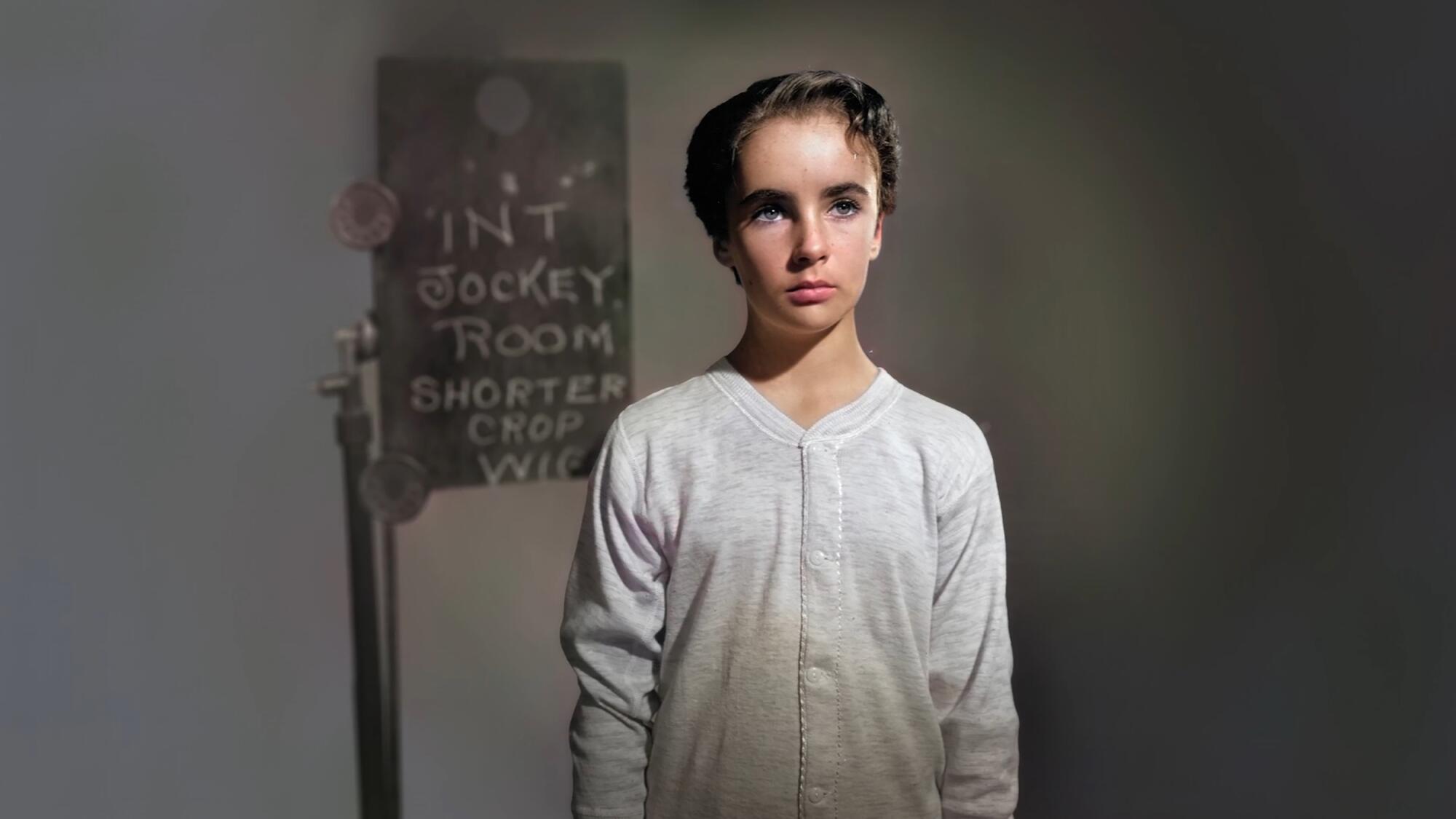 A teenage Elizabeth Taylor stands before a sign that reads "Int Jockey Room Shorter Crop"