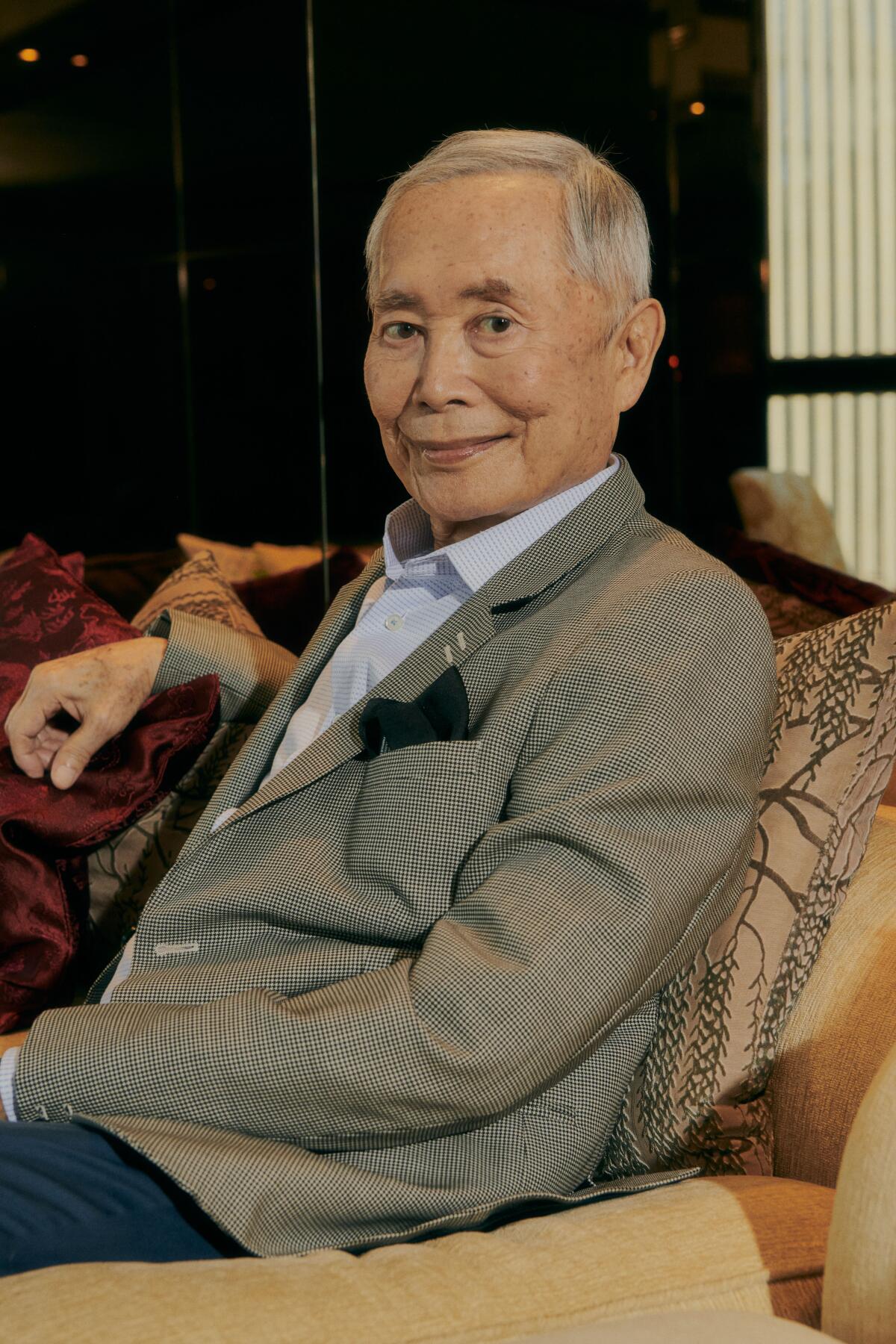 George Takei, in a gray blazer and collared shirt, smiles and leans back into a sofa.